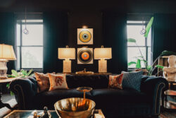 A dark living room with a navy sofa, two table lamps, abstract wall art, and large windows with black curtains.