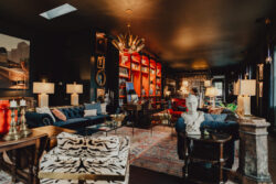 Eclectic lounge with velvet sofas, vibrant shelves, artwork, sculptures, patterned armchairs, and warm ambient lighting.