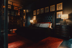 A dimly lit bedroom with a dark bed, red rug, framed art on the wall, and two bedside tables with lamps.