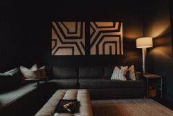 A modern living room with dark walls, a grey sofa, geometric wall art, a lit floor lamp, and a cushioned ottoman.