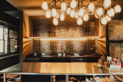 Modern kitchen with metallic backsplash, hanging lights, black cabinets, and various kitchen items on a reflective countertop.