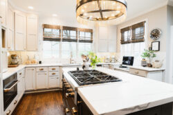 Bright, modern kitchen with white cabinets, gas stove island, pendant light, and large windows with woven shades.