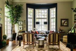 A well-lit living room with large windows, two armchairs, a sofa, lamps, framed art, and decorative plants.