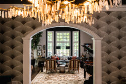 A living room with patterned wallpaper, a chandelier, armchairs, a rug, a grand piano, and large windows.