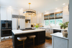 Modern kitchen with white cabinets, black island, gold light fixture, wood floors, and large windows above the sink.