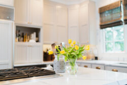 Glass vase with yellow tulips on a white kitchen countertop; cabinets and utensils visible in the background.
