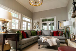 A living room with a gray sectional sofa, colorful cushions, a fur ottoman, wall art, and large windows.