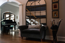 A gray loveseat with a patterned pillow sits by a large mirror and umbrella stand in a hallway near a grand piano.