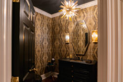 Small bathroom with patterned wallpaper, black vanity, gold fixtures, starburst chandelier, and an oval mirror.