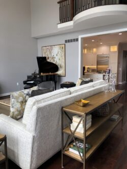 Modern living room with a gray sofa, grand piano, bookshelf, and view into a white kitchen with an island.