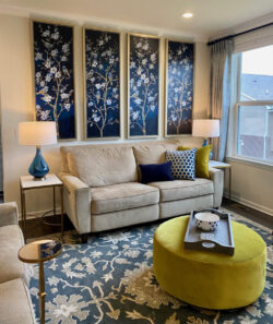 Beige sofa with blue and yellow pillows, yellow ottoman, and floral wall art in a bright, modern living room.