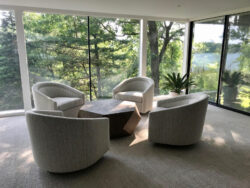Four modern armchairs surround a geometric coffee table in a bright room with floor-to-ceiling windows facing trees.