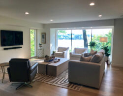 Modern living room with neutral furniture, large windows, and a view of trees and water outside.
