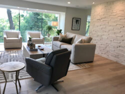 Modern living room with neutral furniture, a textured accent wall, and large windows overlooking greenery.